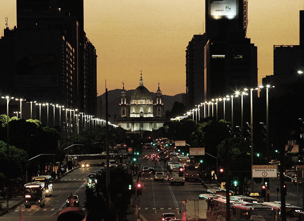 Landscape of a busy avenue, late afternoon. On the sides, several streetlights illuminating the street. Around, buildings surround the image