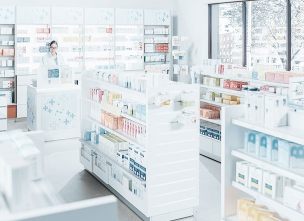 Visão interna de estabelecimento farmacéutico, com gondolas brancas e prateleiras com medicamentos.  