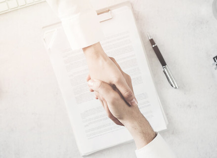 Two white people holding hands as a form of "agreement". In the background, a contract signed by the parties on a table