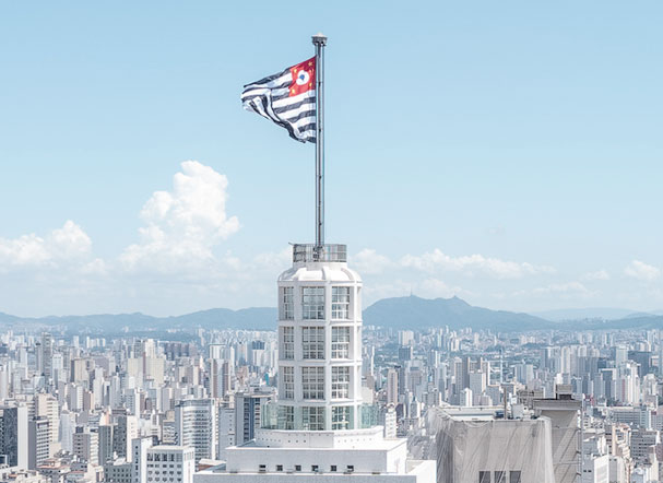 Visão superior de São Paulo, com destaque na bandeira da cidade penturada em uma aste, acima de um edifício